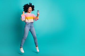 Happy young woman jumping in excitement while holding smartphone, wearing colorful sweater in front...