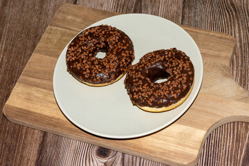 Two delicious chocolate frosted donuts topped with crunchy toffee bits are placed on a pale plate atop a wooden board. The inviting kitchen ambiance enhances their appeal
