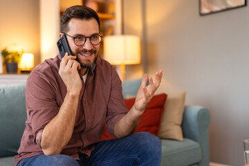 Young man is sitting on a sofa at home, engaged in a phone conversation. The scene captures a...