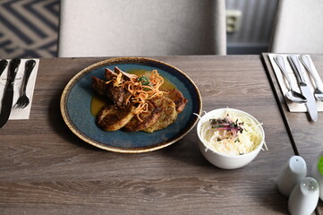 Delicious dish of pork neck with a rich sauce, served alongside crispy potato pancakes. The meal is elegantly presented on a rustic ceramic plate, placed on a wooden restaurant table.
