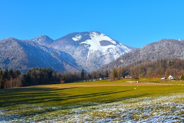 Green field bellow Srednji Vrh snow covered mountain with a forest covered the slopes bellow in...