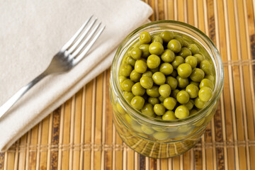 Jar of canned green peas and fork on a tabletop. Open glass jar of marinated sweet peas and metal fork on a table. Vegetable protein, dietary fiber concept.