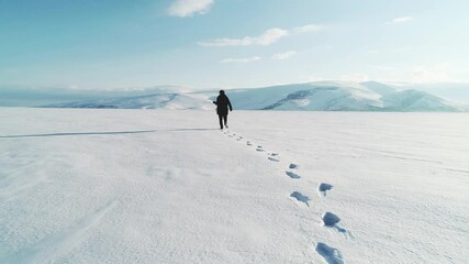 A tourist walks across a vast snow-covered plain, leaving a trail of footprints behind. Distant mountains rise under a clear blue sky. The untouched snow stretches endlessly in every direction.