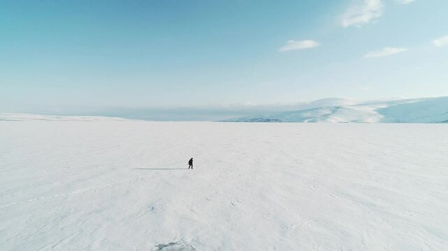 A tourist walks across a vast snow-covered plain, leaving a trail of footprints behind. Distant mountains rise under a clear blue sky. The untouched snow stretches endlessly in every direction.