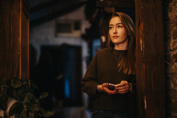 A young woman stands indoors holding a smart phone, gazing with a thoughtful expression amidst a...