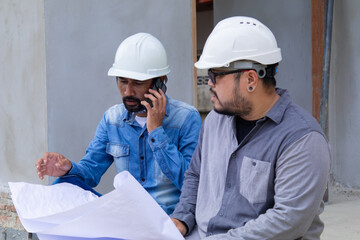 Two construction engineers in hard hats serious discussion over blueprints at construction site, Asian architect talking to foreman standing at unfinished building structure in real estate investment
