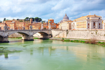 Scenic view of Ponte Vittorio Emanuele over Tiber River, with St. Peter Basilica rising in background in Rome Italy. Historic architecture, dramatic clouds, and reflections create timeless atmosphere