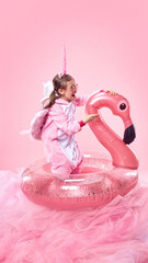 Obraz premium Girl in pink unicorn onesie excitedly playing with inflatable pink flamingo against pink background. Summertime vacation vibes. Concept of childhood, fantasy, dreams, imagination