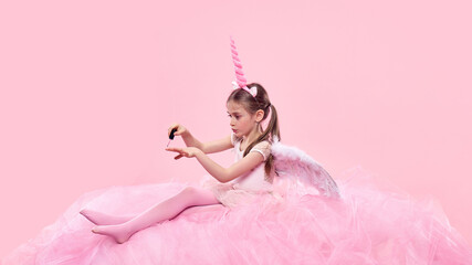 Girl in pink fairy costume with unicorn horn carefully painting nails, sitting on pink fabric against pink background. Taking care, beauty salon. Concept of childhood, fantasy, dreams, imagination © master1305