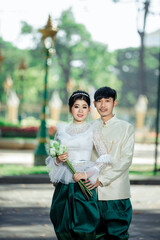 A young Cambodian couple in traditional wedding attire posing outdoors in a park with golden ornaments and lush greenery