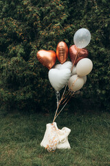 A stylish bouquet of balloons in shades of white, silver, and copper against a green tree backdrop tied to a bag. A decoration for parties, weddings, anniversaries, birthdays, baby showers.