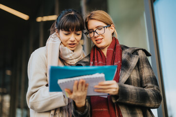 Two women engage in a collaborative discussion while reviewing documents outside a modern building.