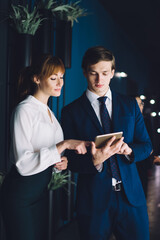 Pensive business partners using digital tablet while multiethnic coworkers having conversation in lobby of modern office building