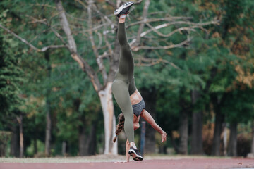 A fit woman performs a handstand on a track in a scenic, lush park, showcasing her strength,...