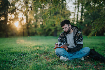 Obraz premium A man sits on the grass writing in a notebook, surrounded by lush greenery and a beautiful sunset. The peaceful setting enhances creativity and reflection in this tranquil outdoor scene.