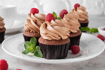 Chocolate cupcakes with cream decorated with fresh raspberries and mint leaves on a gray concrete background. Delicious holiday dessert