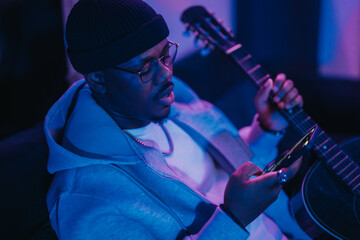 Obraz premium Musician intently uses a smart phone while holding a guitar in a recording studio. The creative ambiance is highlighted by dim, colorful lighting, symbolizing focus, inspiration, and musical creation.
