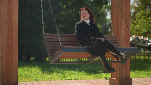 Stylish woman in black coat and boots serenely sits on wooden swing, hands clasped between legs, gazing into the distance in a sunlit par