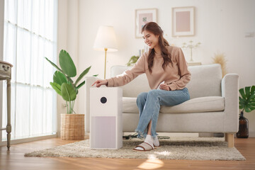 Healthy Asian woman is operating an air purifier while relaxing at home, enjoying a moment of...