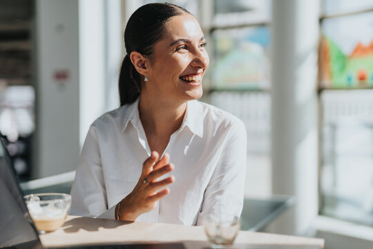 A smiling businesswoman takes a coffee break in a bright, modern office kitchen. She appears relaxed and content, enjoying a moment away from work. Sunlight streams through the large windows.