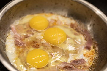 Close-up of three sunny-side up eggs frying in a pan with bacon.