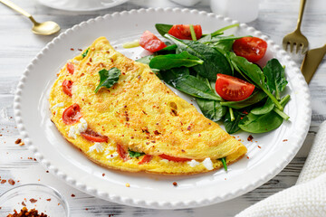 Stuffed omelette with tomatoes and ricotta cheese, served with fresh spinach leaves and cherry tomatoes. Healthy breakfast. White wooden background