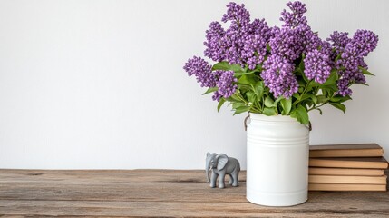 Fototapeta premium Purple lilac flowers in a white milk can sit on a rustic table with books and an elephant figurine against a white wall