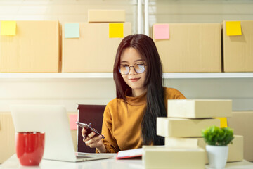 Asian owner holding a smartphone. Female entrepreneur preparing orders for shipping in a warehouse....