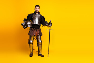 Naklejka premium Man in full knight armor holding sword and helmet isolated on yellow background showcasing historical medieval theme