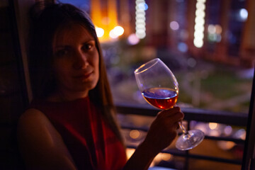 A girl drinks with a glass near the window in the evening looking at the city lights
