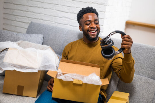 Happy young African-American man smiling and opening delivered package with headphones while sitting on sofa at his home. Image captures joy and convenience of home shopping and receiving order.
