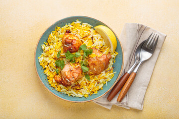 Delicious indian chicken biryani with chicken, garnished with fresh cilantro and lemon, in a bowl on a beige table, accompanied by cutlery. Authentic dish of India