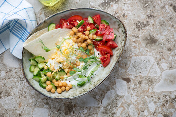 Yogurt bowl with vegetables, chickpeas, grated egg and flat bread, horizontal shot on a light-brown granite background with space, above view