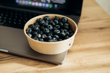 A laptop on the table and a bowl of fresh blueberries as a snack.