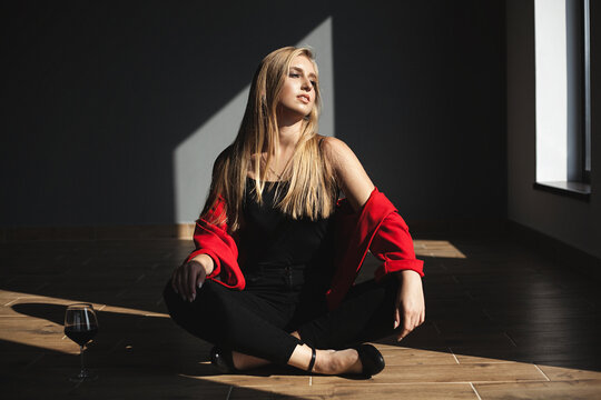 a woman with long blond hair sits on the floor in the shade. She is wearing a black top and black trousers, and has a red jacket or blazer over her shoulders. - Powered by Adobe