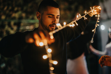 A young man is arranging glowing string lights, creating a warm and festive atmosphere in a dimly...
