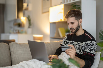 Man shopping online with a laptop and credit card while relaxing on a sofa in a cozy living room