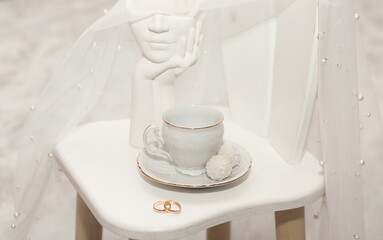 Elegant cup with sweets and wedding rings on white background