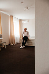 A man in a white shirt and dark trousers stands leaning on the headboard of a bed in a bright room. He is wearing white sneakers and wears glasses.