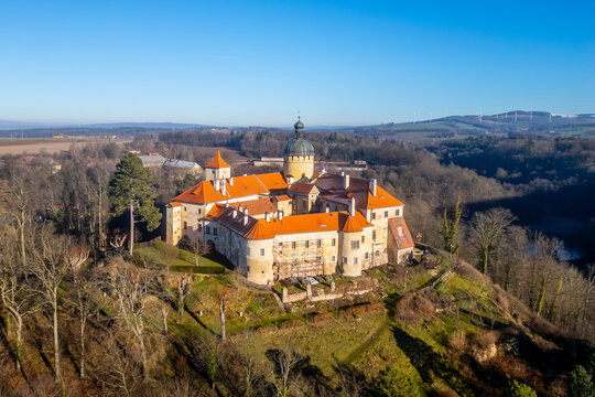 Grabstejn castle stands majestically on a hill, surrounded by lush greenery. The clear blue sky highlights the castle's vibrant orange roof and intricate architecture, creating a picturesque scene.