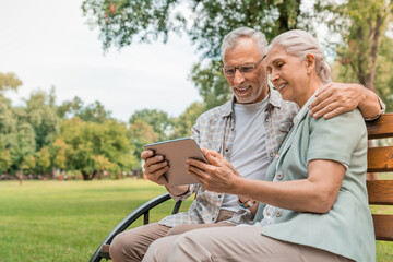 Caucasian senior elder couple sit on bench in park. Mature happy and enjoy with slow life. Old man and woman look at tablet and smile with life together. Retirement warm family lifestyle concept.