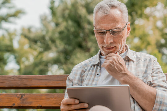 Smiling modern old man in casual eyeglasses relaxing city park. Pensive senior gray haired male sitting wooden bench outdoors using holding digital wireless tablet pad browsing internet watching video