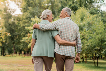 Rear view of senior couple walking outdoors in summer park and embracing, loving happy mature spouses hugging and smiling to each other, cheerful husband and wife enjoying outside walk, copy space