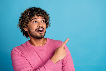 Portrait of a cheerful young man with curly hair in a pink sweater pointing upward against a solid...