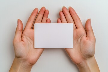 Fototapeta premium A pair of hands holding and presenting an empty white card, isolated on the left side against a plain background. The focus is on capturing both palms with their fingers gently pinching the
