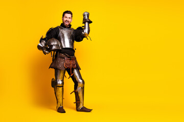 Young man in shiny medieval armor holding a helmet with a cheerful gesture on vibrant yellow...