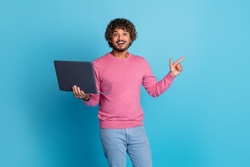 Smiling young man with curly hair in a pink sweater holding a laptop, gesturing energetically...