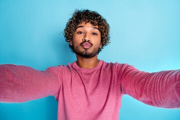 Trendy young man in casual pink sweater posing for a selfie with a fun expression, set against a vibrant blue background