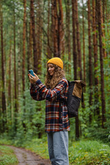 Young female hiker wearing a yellow beanie and carrying a backpack, using a smartphone to capture pictures while walking along a scenic forest trail during summer vacation