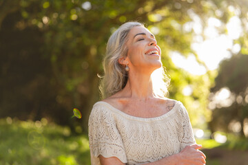 A joyful mature woman basks in the sunlight, relishing the surrounding nature in a vibrant, lush...